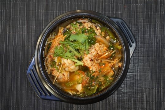 위에서 내려다본 뚝배기 탕 — 주황빛 국물, 알 덩어리 같은 건더기, 흰 주름진 건더기, 푸른 잎채소, 팽이버섯; 남색 받침, 짙은 회색 매트